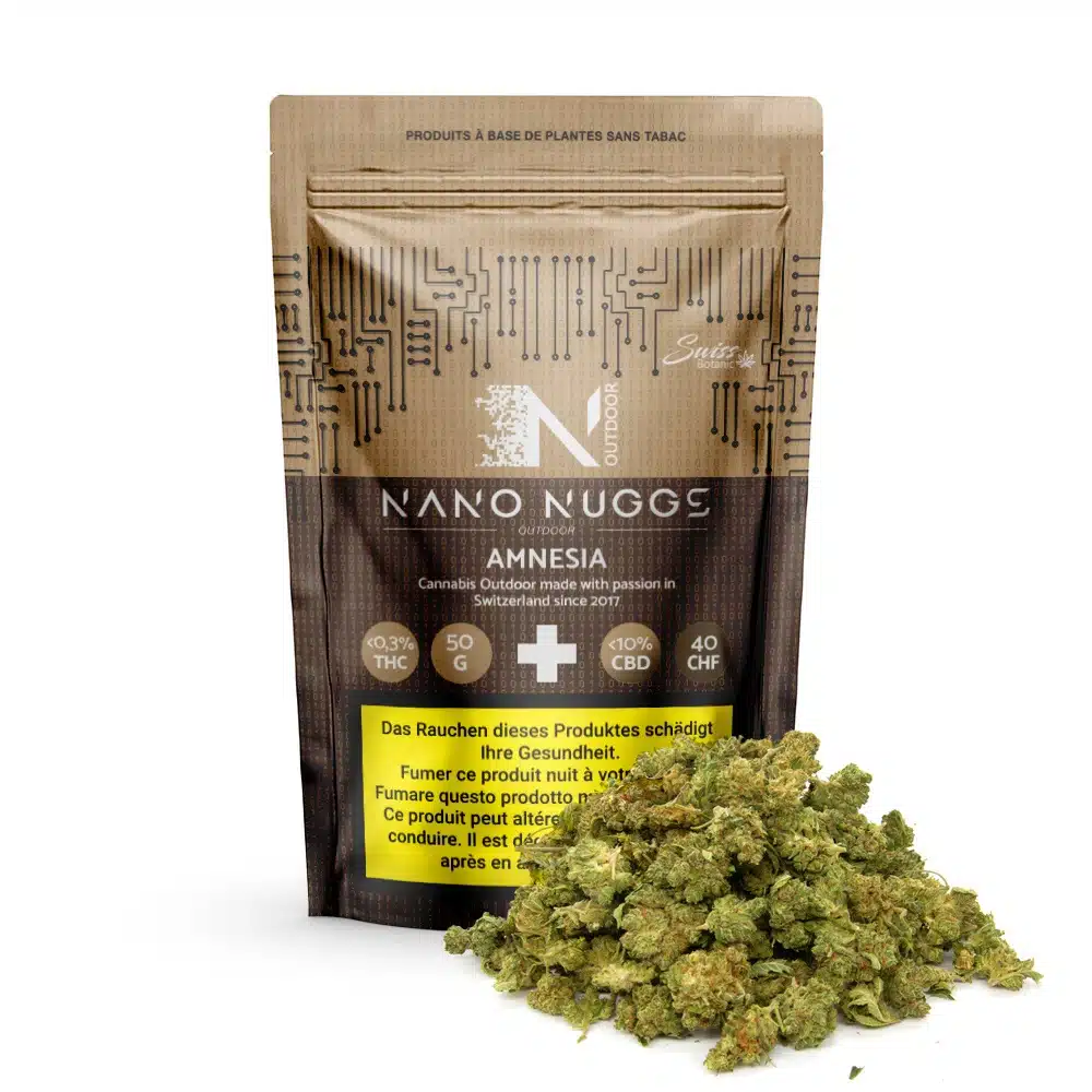 Fleurs de cbd outdoor nano nuggs - amnesia – 0. 3% thc 1 Un sachet brun refermable étiqueté "fleurs de cbd outdoor nano nuggs - amnesia - 0,3% thc" se trouve derrière une pile de bourgeons de cannabis nano nuggs, affichant les détails du produit et une étiquette jaune d'avertissement sanitaire sur le devant.