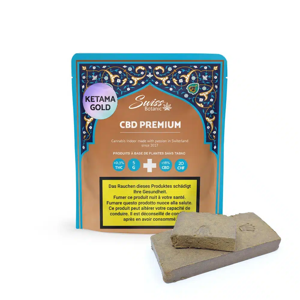 A package of Ketama Gold Hash CBD Premium stands behind two brown hashish blocks, displaying product details and health warnings in several languages on the packaging.