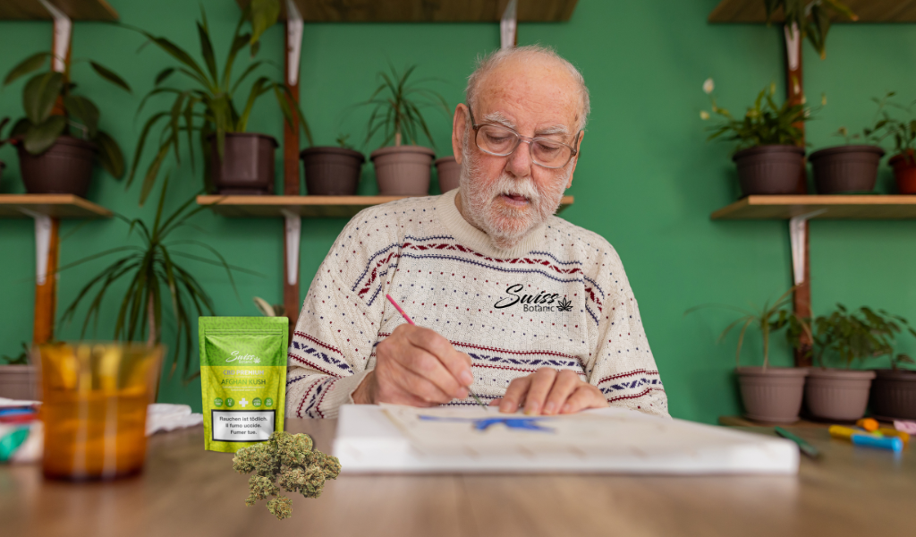 An elderly man with glasses is focused on writing in a notebook at a table with cannabis products and plants nearby.