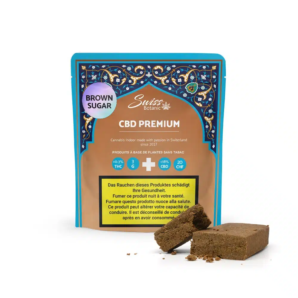 A package labeled "Brown Sugar Hash CBD Premium - 0.3% THC" with a decorative design contains cannabis resin resembling brown sugar, with several resin pieces displayed in front of the package.
